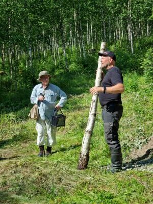 Mies seisoo loivassa rinteessä ja pitää parimetristä visakoivurunkoa nojallaan itseään vasten. Toinen mies kuuntelee hänen puhettaan. Heidän takanaan kasvaa nuoria koivuja rehevässä vihreässä maapohjassa. 
