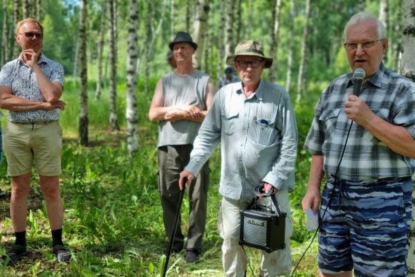 Koivumetsässä seisoo neljä miestä, joista oikealla oleva puhuu mikrofoniin. Hänen vieressään seisova mies kantaa suurta äänenvahvistinta. 