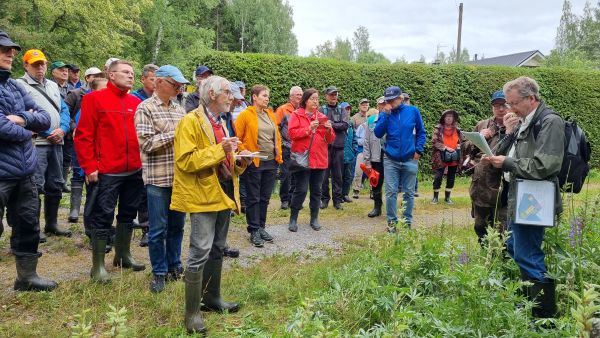 Havupuuaidan edessä seisoo kirjava joukko miehiä ja naisia, jotka kuuntelevat karttalaukku olallaan seisovaa miestä. 