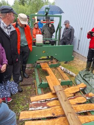 Valkoseinäisen rakennuksen vieressä on vihreä piensaha, jonka linjalla on useita sahattuja visakoivupätkiä. Sahalaitteen ympärillä on useita ihmisiä katselemassa sitä. 