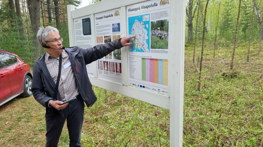 Silmälasipäinen mies osoittaa sormellaan ulkona olevaa opastaulua ja siinä olevaa karttakuvaa. 