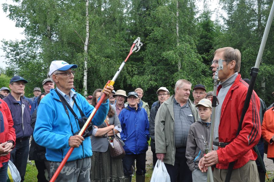 Taustalla on suureksi kasvanutta koivikkoa. Sen edessä on tiealue, jolla seisoo miehinen ryhmä. Etualalla on vastakkain kaksi miestä, joilla molemmilla on pitkävartisia metallisia oksaleikkureita. 
