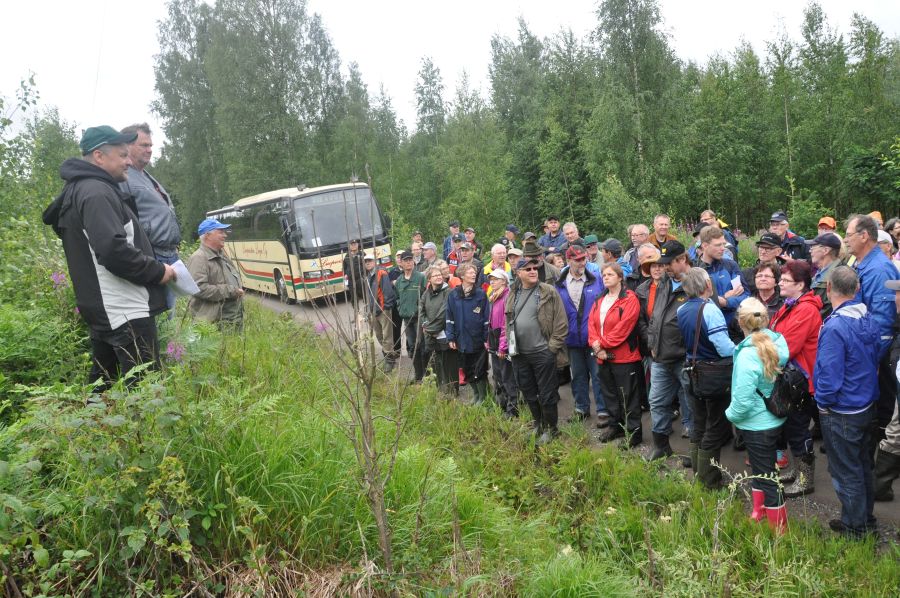 Hiekkatiellä on suuri keltasävyinen linja-auto. Sen edessä on suuri joukko ihmisiä kuuntelemassa edessään olevaa mieskolmikkoa, joka seisoo vähän korkeammalla. heinikkoisella pientareella