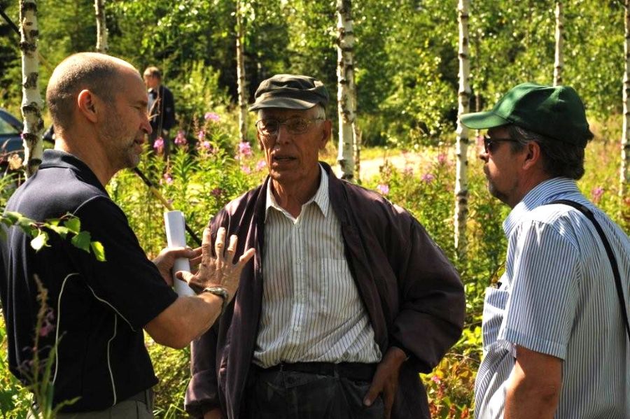 Yksi paljaspäinen ja kaksi lippalakkipäistä miestä keskustelee nuoren, muhkurarunkoisen ja vaaleatuohisen koivikon edessä. 