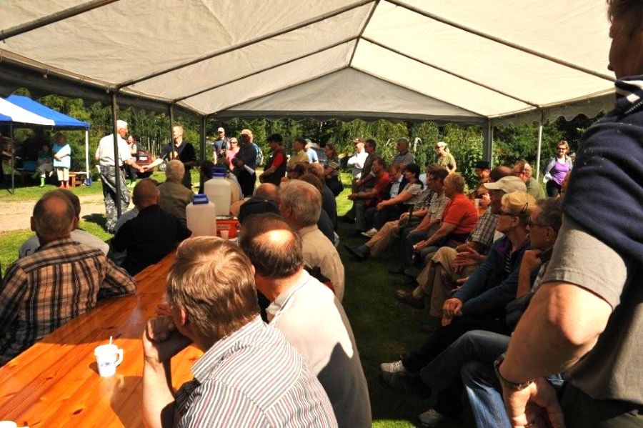 Valkoisen pitkänomaisen katoksen alla on pitkiä pöytä ja sen ääressä runsaasti miehiä. Pöydällä on vesikanistereita ja kertakäyttömuki. Taustan pihanurmikolla kaksi miestä kättelee toisiaan. 