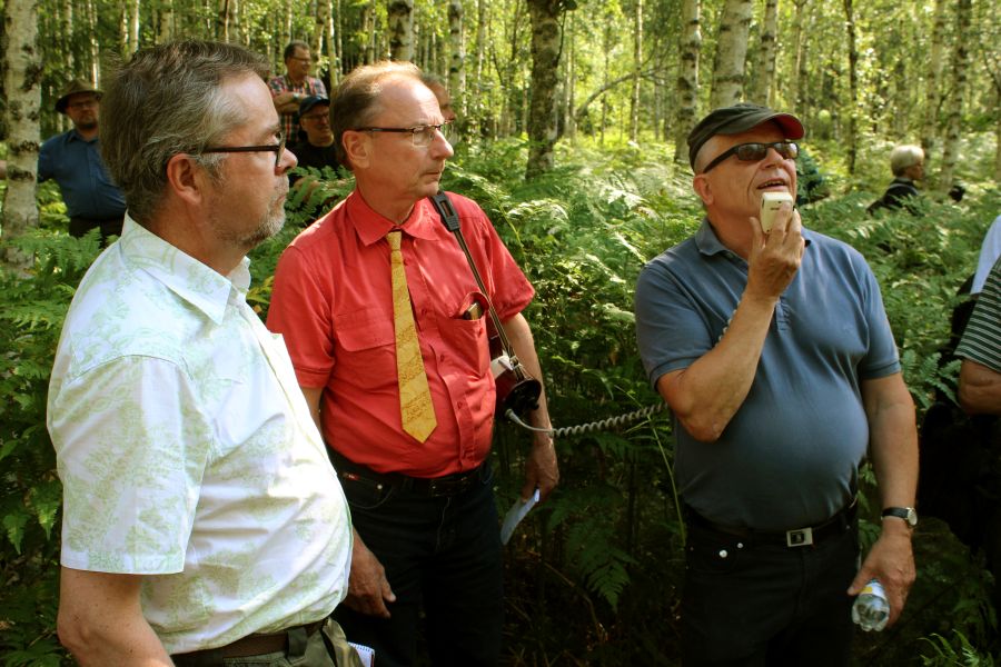 Kolme silmälasipäistä miestä, pukeutuneina valkoiseen, punaiseen ja siniseen paitaan, seisovat sananjalkojen ympäröiminä nuoressa koivikossa. Punapaitaisella miehellä on visakoivuinen kravatti. 