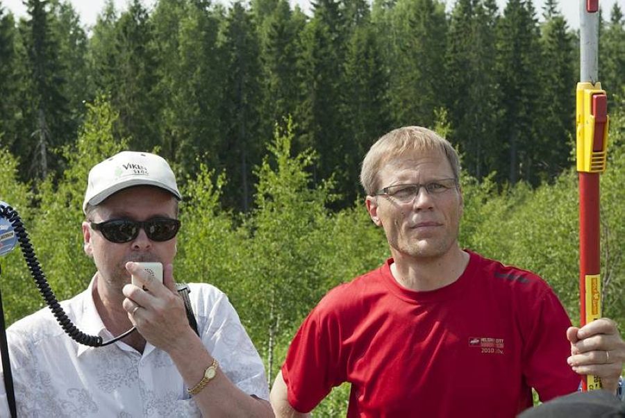 Kaksi miestä seisoo matalan koivikon edessä. Koivikon takana on korkea kuusikko. Vasemmalla seisova, valkoasuinen mies puhuu mikrofoniin. Punapaitaisella on pitkävartinen oksasaha.