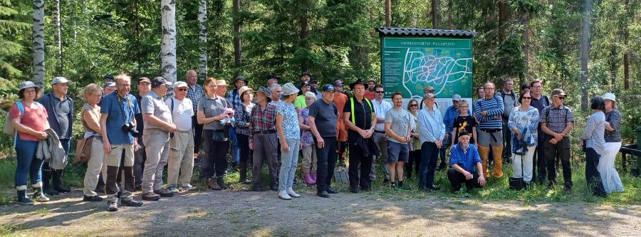 Suuri joukko kesäasuisia miehiä ja naisia seisoo hiekkatiellä metsän edessä. Heidän vieressään on suuri kyltti, jossa on kartta ja lukee: Hanneksen metsä – Puulajipuisto. 
 
