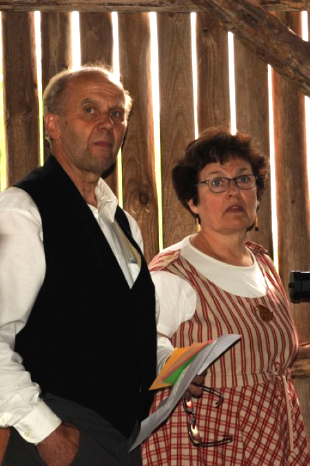 Kansallispukuiset mies ja nainen seisovat harvalaudoitetussa ladossa. 