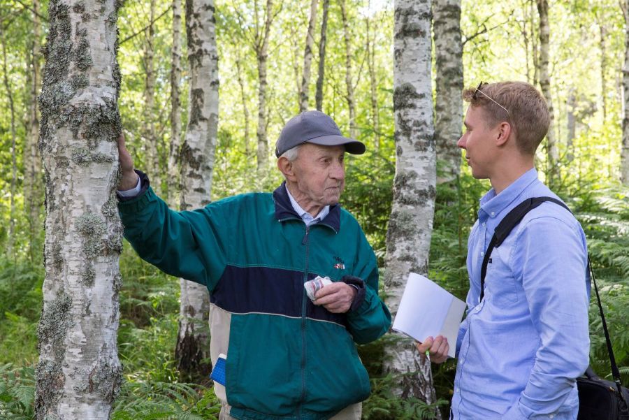 Iäkäs mies on vastatusten nuoren miehen kanssa koivikossa. Nuorella miehellä on kädessään lehtiö. 