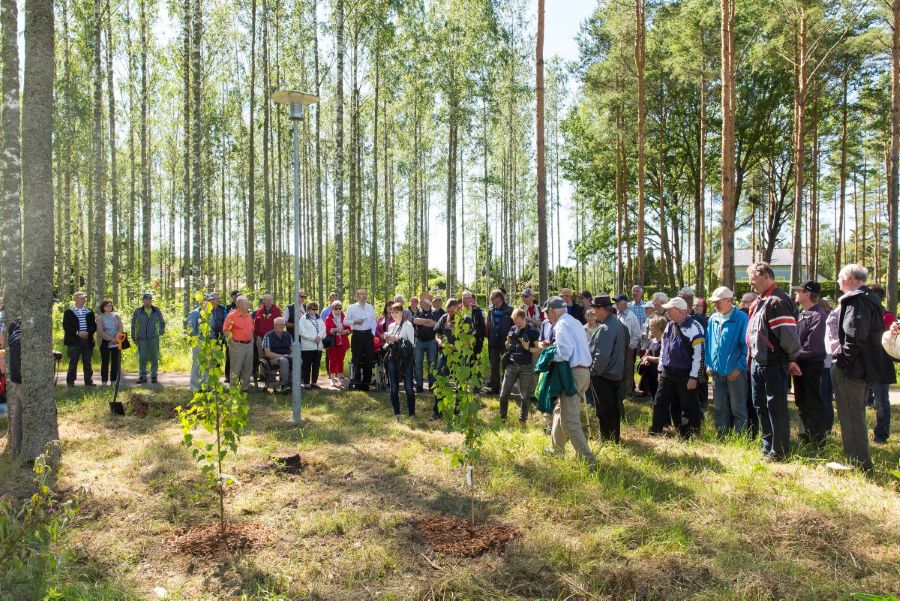 : Iso joukko ulkoasuihin pukeutuneita ihmisiä seisoo metsän keskellä olevalla heinittyneellä aukiolla. Takana on koivikko ja männikkö. Aukiolla on kaksi pientä vihreälehtistä koivua, joiden tyvellä näkyy paljastettua maata. 