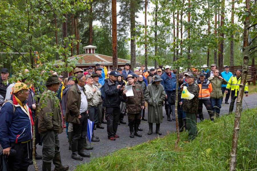 Sadeasuinen miesjoukko kuuntelee ulkona lippalakkista miestä, joka puhuu mikrofoniin. Miesten takana näkyy muutaman rakennuksen katot.