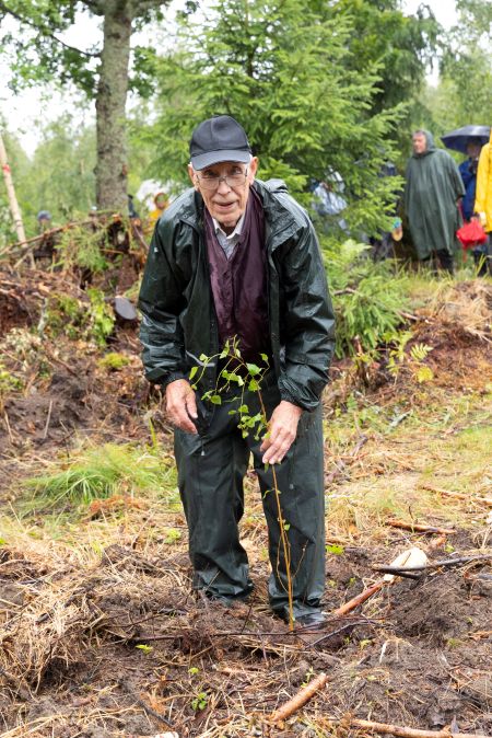 Tummaan sadeasuun pukeutunut lippalakkipäinen mies pitää vasemmalla kädellä kiinni vasta istuttamansa koivuntaimen yläosasta ja katsoo samalla kameraan.

